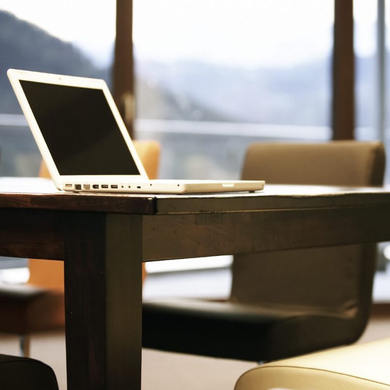 Ein Schreibtisch mit einem offenen Laptop und Stühlen im Hintergrund.